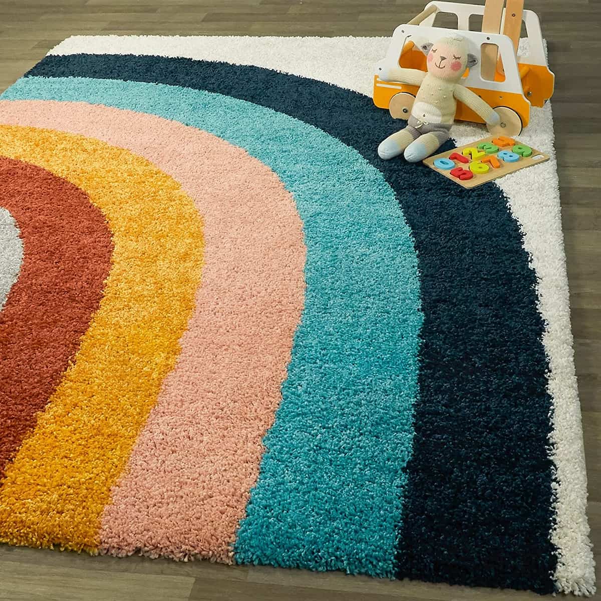 Carpet Mahal's multicolored microfiber shaggy rug with a semi-circular pattern in shades of white, blue, navy, pink, gold, red, and grey, placed on a wooden floor with a toy car and a doll on top of it.